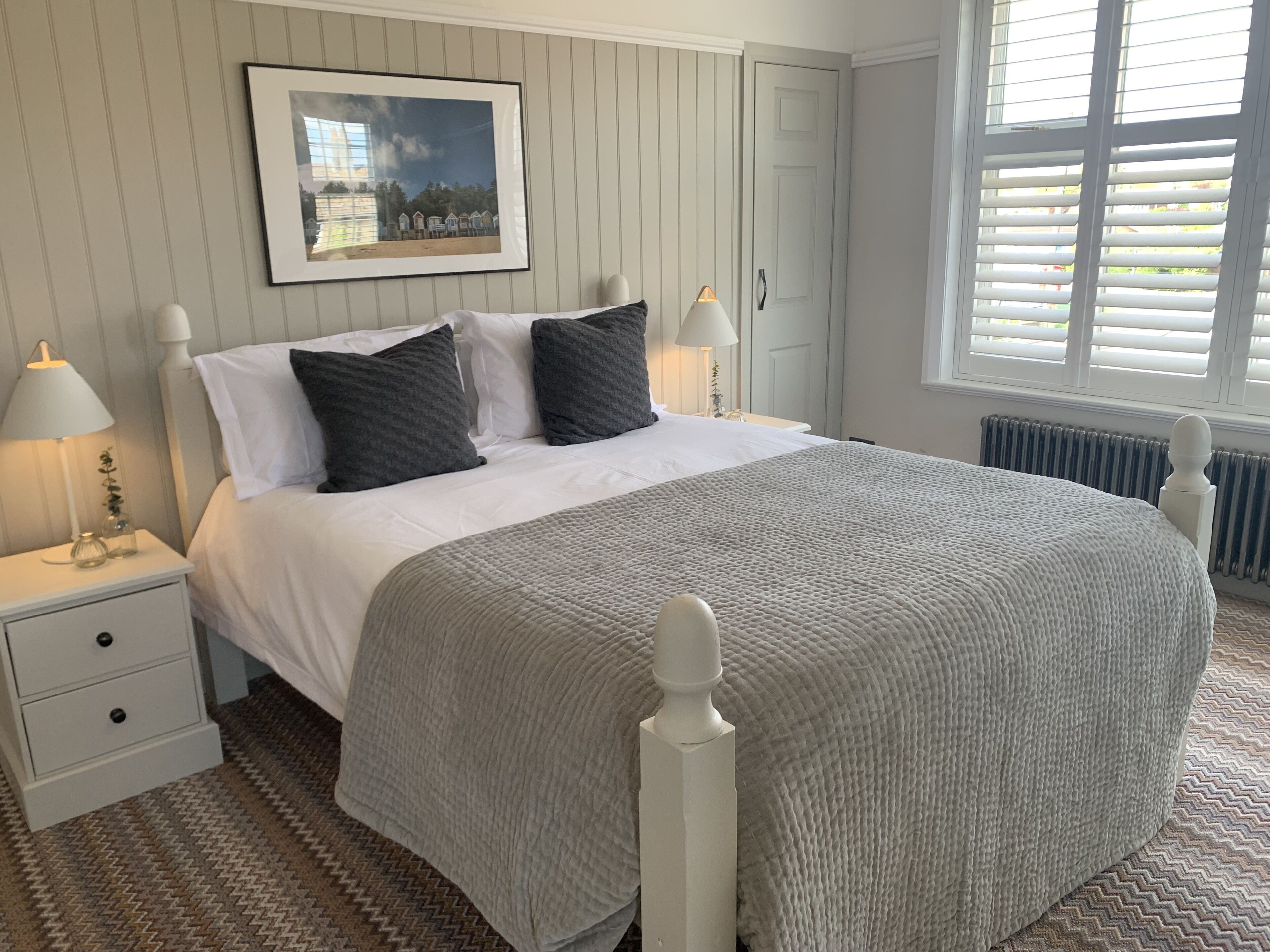 Een netjes ingerichte slaapkamer met een bed, nachtlampjes, een lijst met een foto aan de muur en een raam met luxaflex.