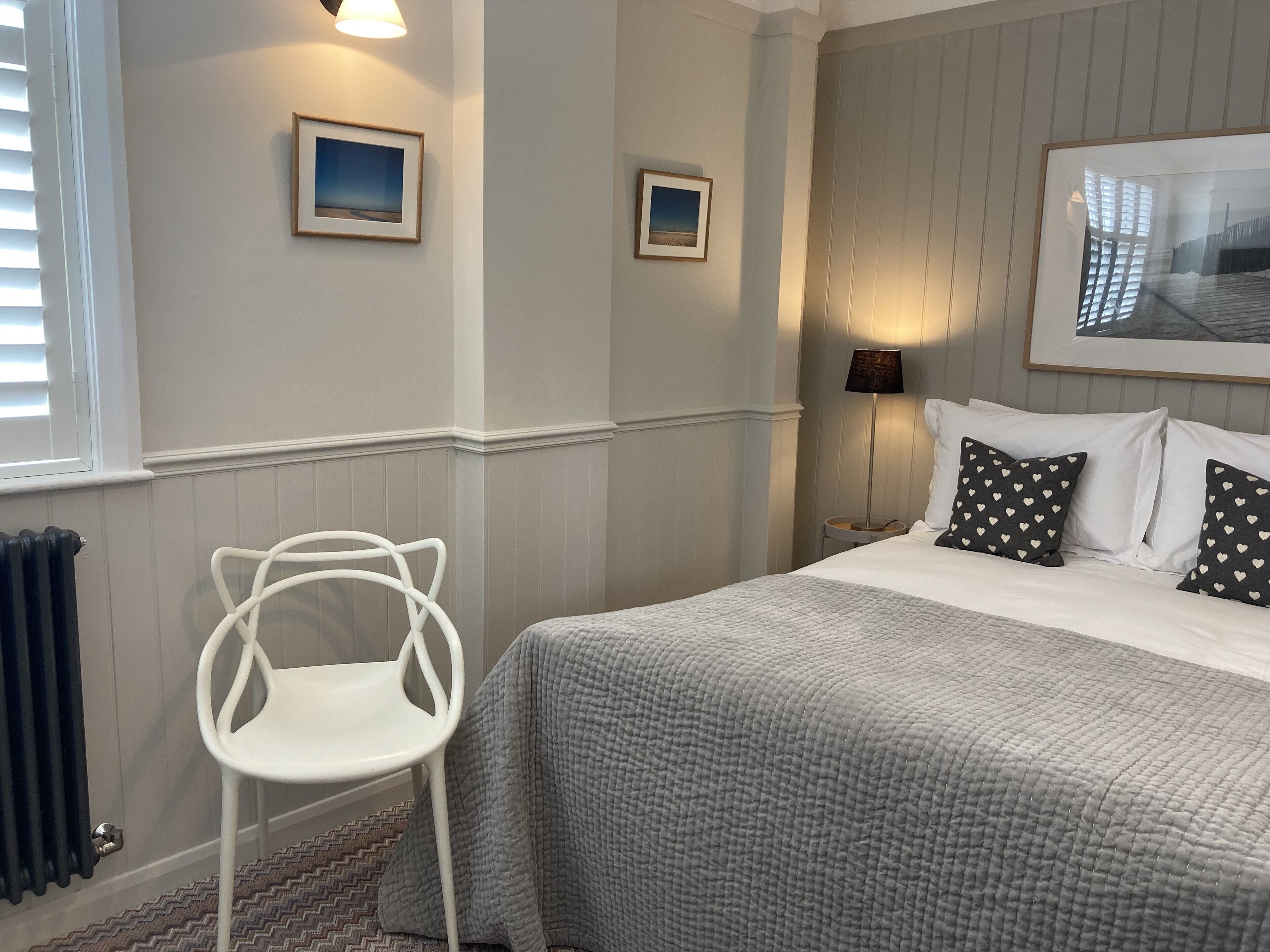 A neatly arranged bedroom with a bed, two framed pictures on the wall, a standing lamp, and a white chair.