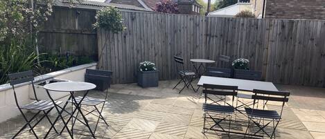 An outdoor patio area with a wooden fence, a brick house in the background, and a tiled floor.