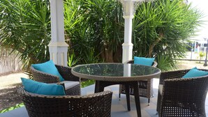 A patio area with wicker furniture and a glass table, surrounded by lush greenery. - Ti Cocon de Standing (Saint-François)