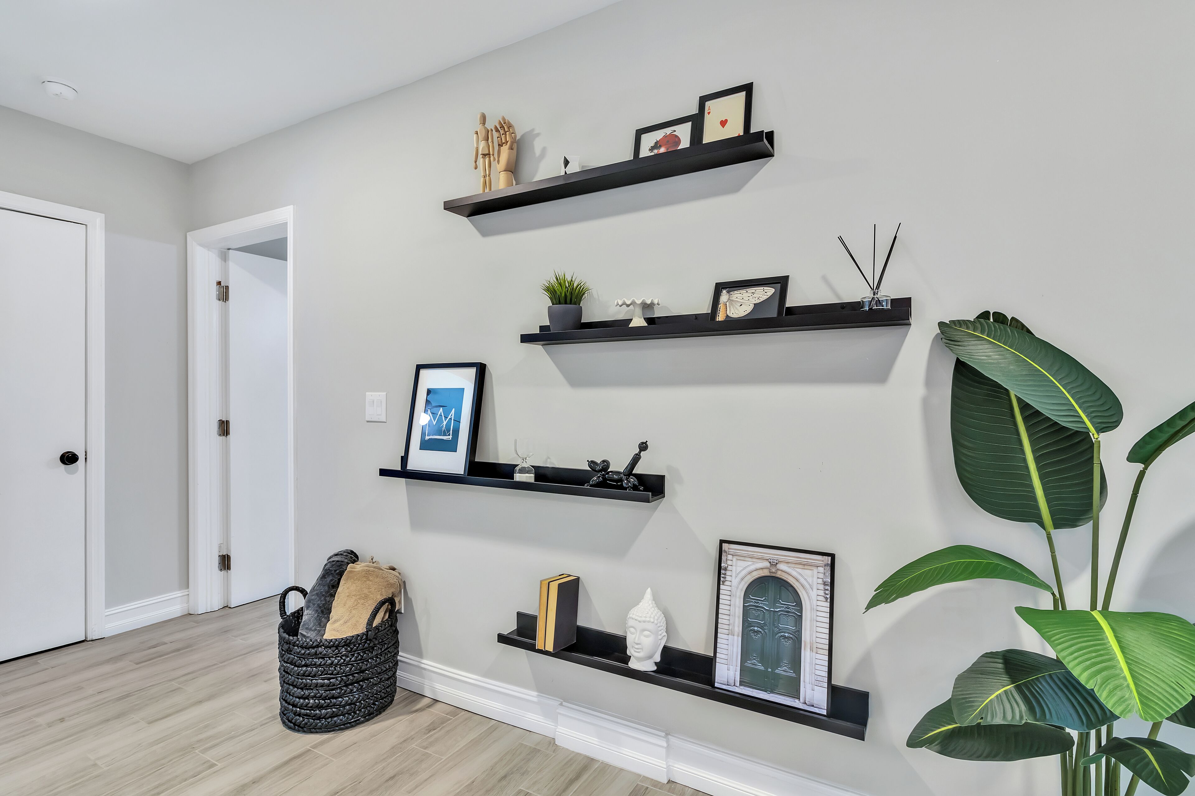 A hallway with three black wall-mounted shelves displaying various decorative items, a woven basket with a beige cushion, and a large green plant.