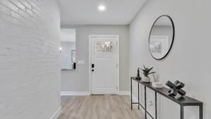 Un couloir avec un mur en briques blanches, un plancher en bois, une porte blanche, un miroir à cadre noir et une console sur laquelle sont posés un pot de plantes et des objets décoratifs.