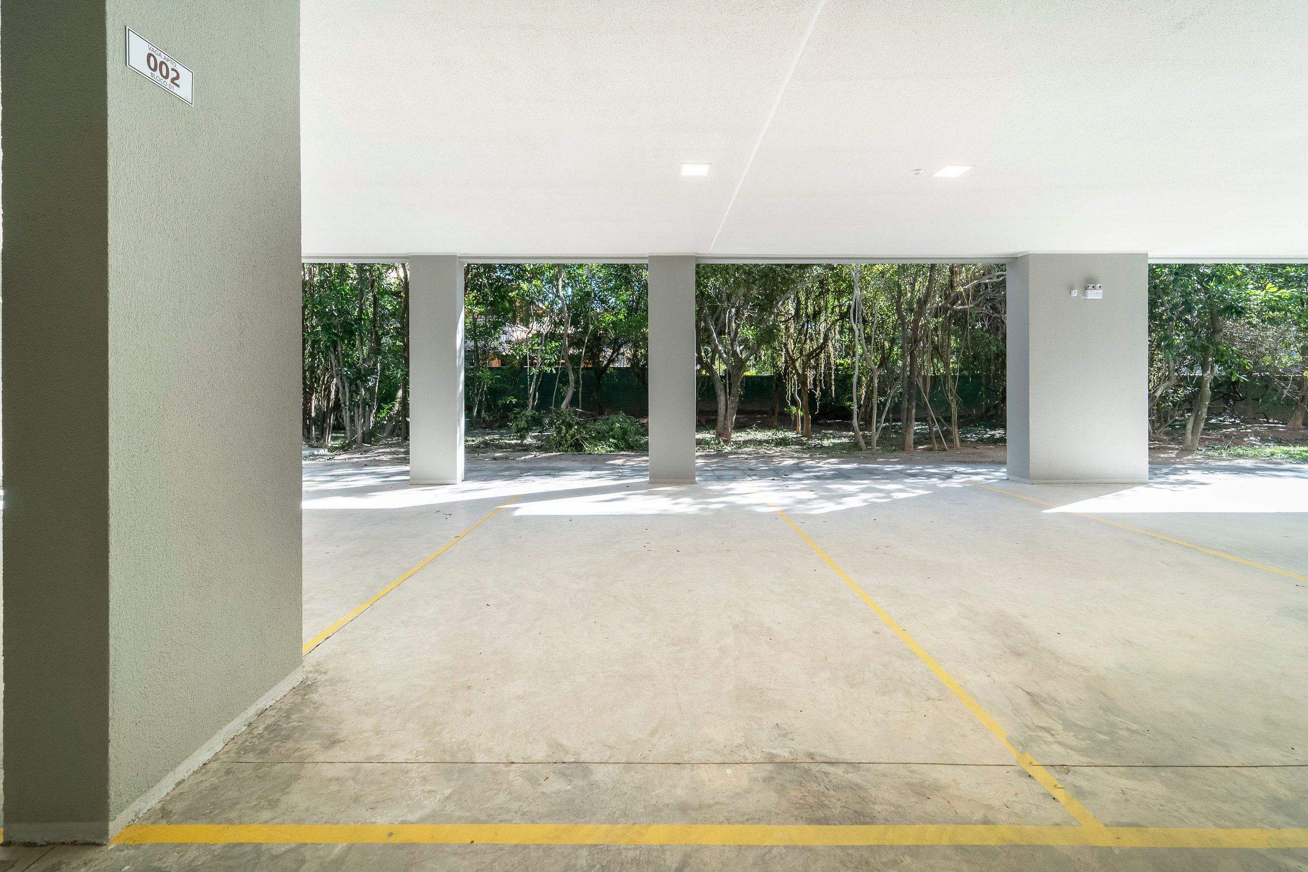 An empty parking area with concrete floors and yellow lines, surrounded by trees and a white wall with a number sign.