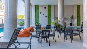 A modern outdoor seating area with a pool, featuring a mix of tables and chairs, some with orange cushions, and potted plants. - 864C Duplex 80 mts Mar Canto Grande SC (Bombinhas)