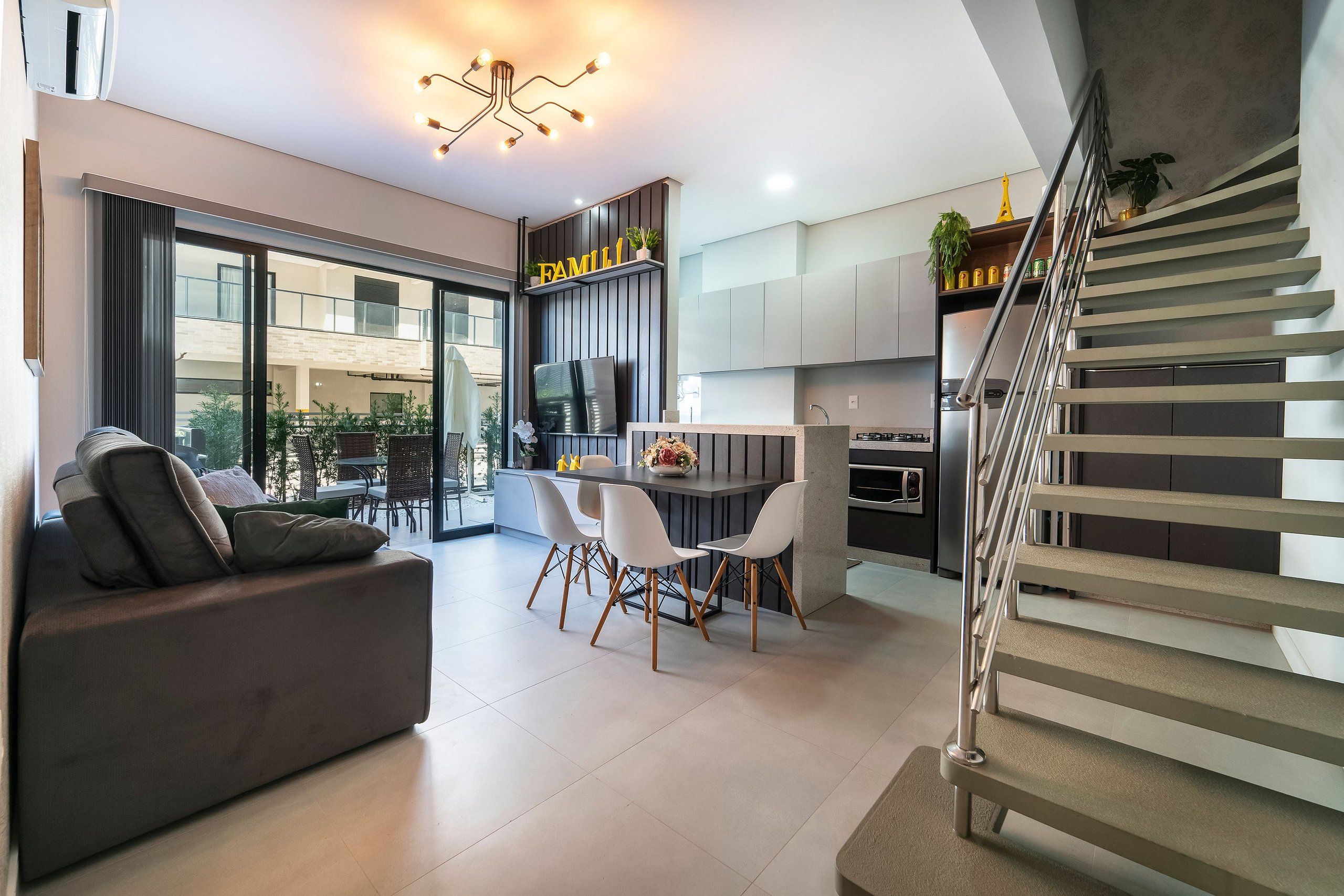 A modern living room with a sofa, dining table, and chairs. There is a kitchen area with cabinets and a staircase.
