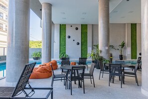Un área de estar moderna al aire libre con piscina, que cuenta con una variedad de mesas y sillas, algunas con cojines anaranjados, y plantas en macetas.