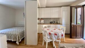 A compact room with a bed, a dining table with chairs, a kitchenette, and a sliding door to a balcony. - Appartamento Il Bottaio (Domodossola)