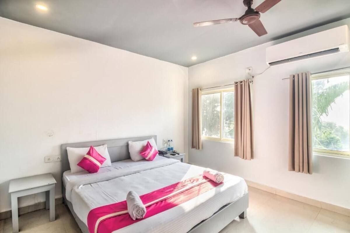 A hotel room with a bed, a nightstand, a ceiling fan, an air conditioning unit, and a window with curtains.