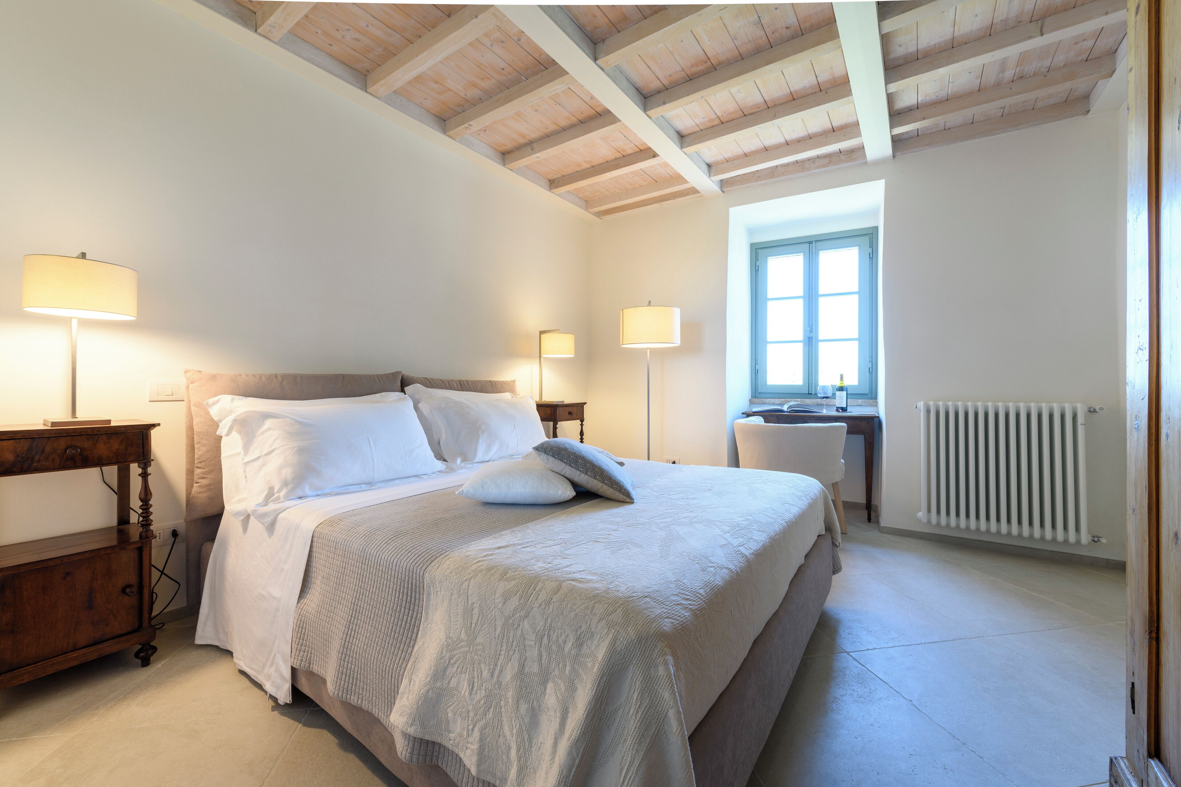 Un dormitorio con una cama grande, lámparas de noche, cabecero de madera, una ventana con persianas y un radiador.