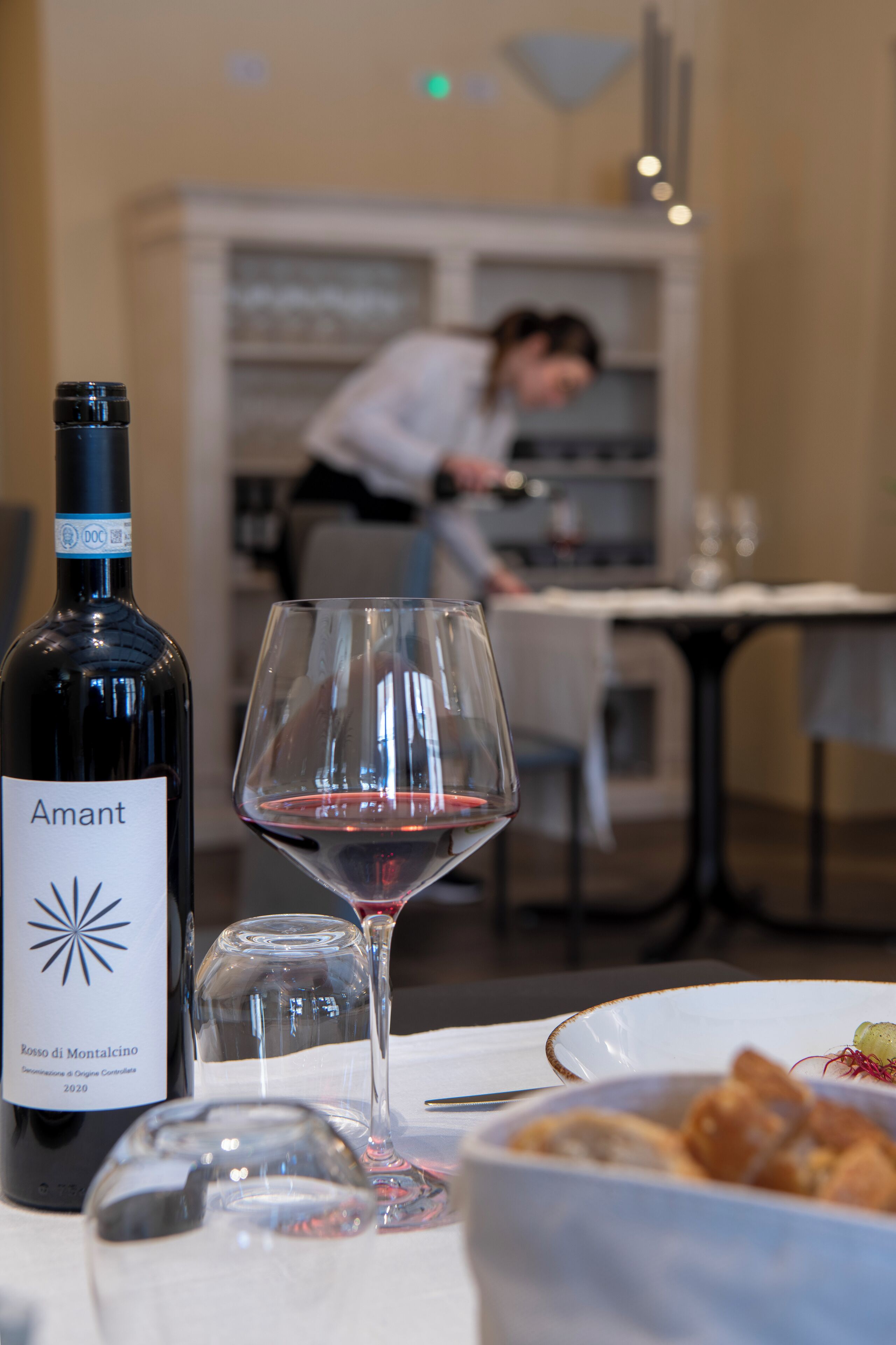 A bottle of wine and a glass on a table, with a bowl of food and a person in the background.