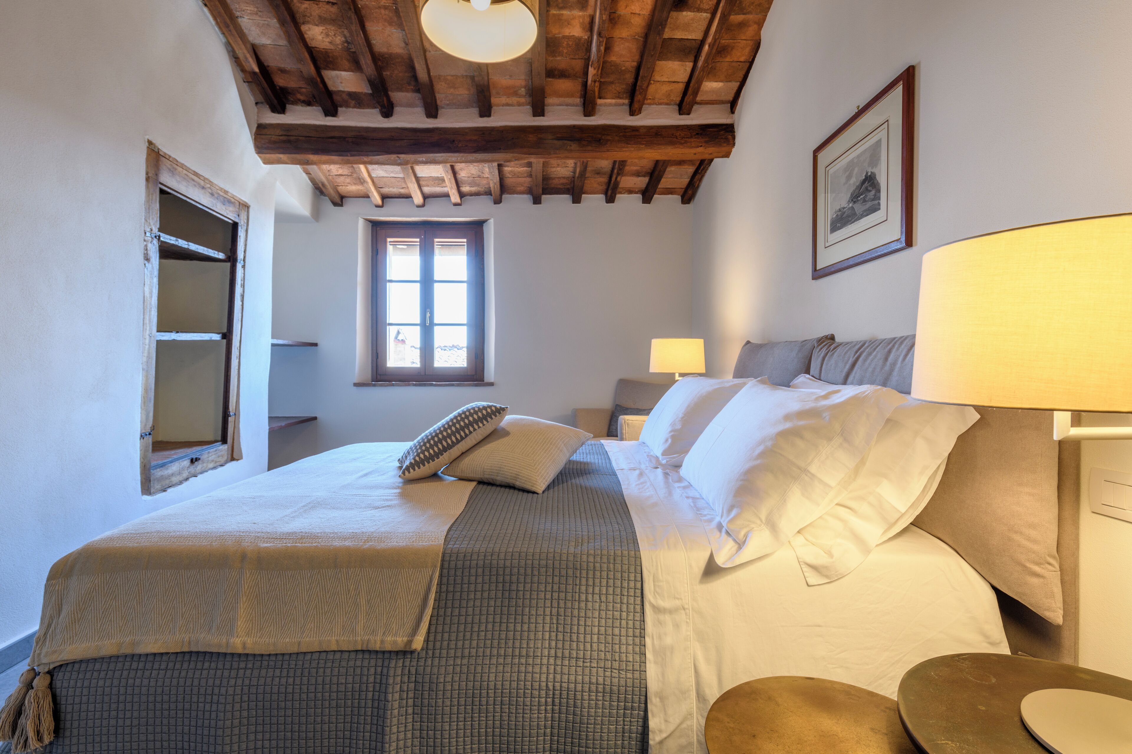 Un dormitorio con techo de madera, una cama grande con ropa blanca, una ventana y una mesita de noche con lámpara.