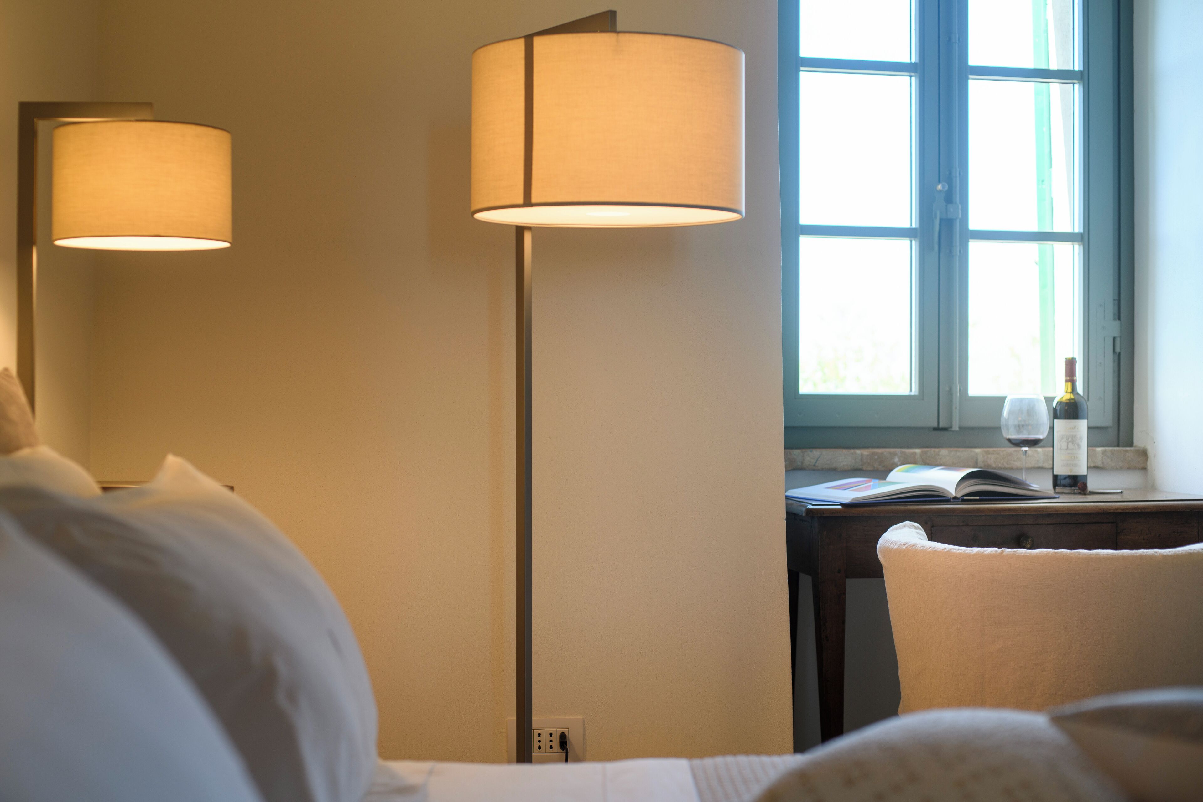 Ein Hotelzimmer mit einem Bett, einem Schreibtisch mit einem Buch und einer Weinflasche, einer Lampe und einem Fenster mit Tageslicht.