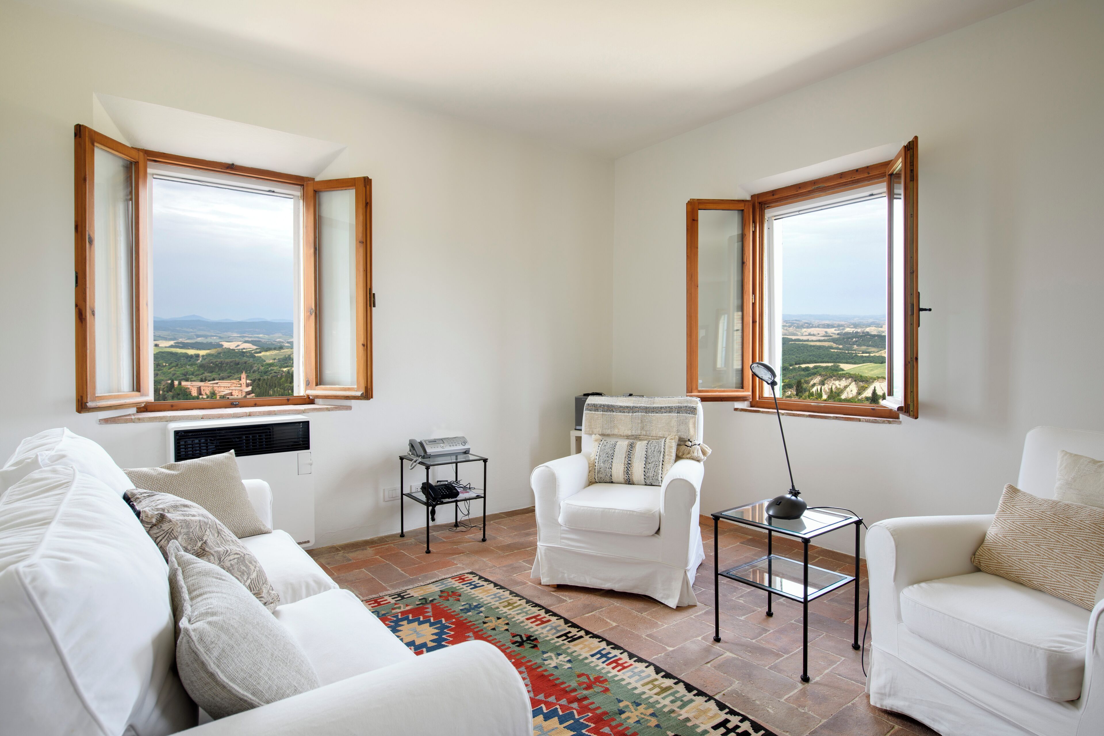 Un salon moderne avec un canapé blanc, un fauteuil blanc et une petite table sur laquelle se trouve une lampe. Il y a deux fenêtres à cadres en bois, dont l’une offre une vue sur un paysage.