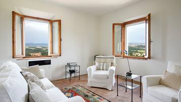 Un salon moderne avec un canapé blanc, un fauteuil blanc et une petite table sur laquelle se trouve une lampe. Il y a deux fenêtres à cadres en bois, dont l’une offre une vue sur un paysage.