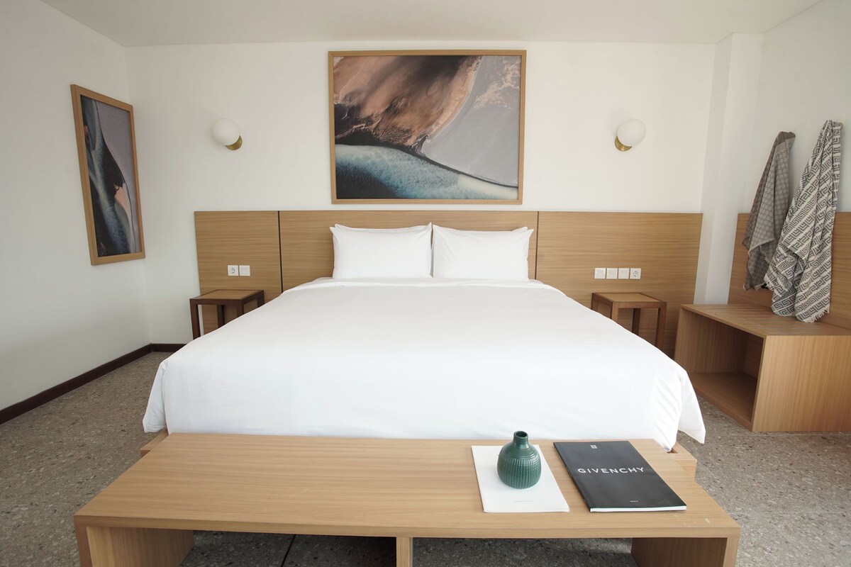 Ett hotellrum med en stor säng, ett träbord vid sängen, ett litet bord med en bok och en vas, och en tavla på väggen.