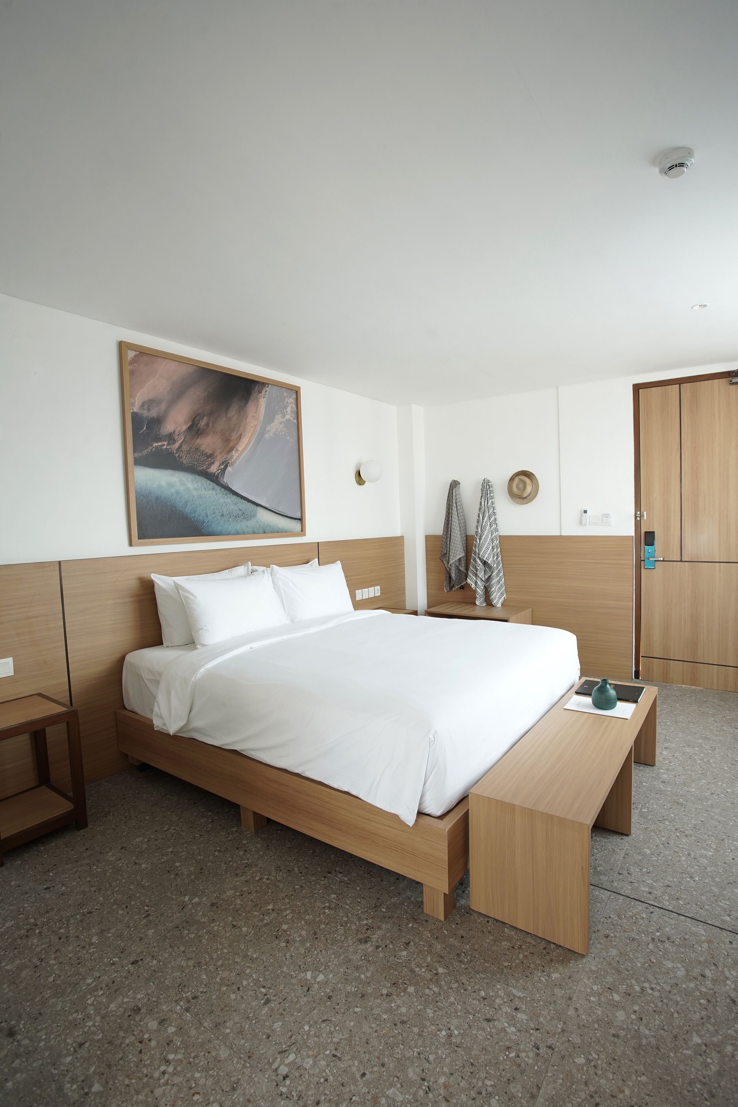 A modern hotel room with a large bed, wooden headboard, a bench at the foot of the bed, a nightstand, a wall-mounted hat rack, and a wooden wardrobe.