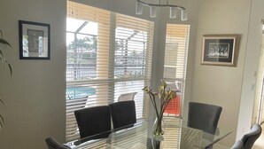 A dining area with a glass table surrounded by four chairs, a vase with flowers, and two framed pictures on the walls. - Villa Meridian Spacious villa for discerning guests. (Cape Coral)