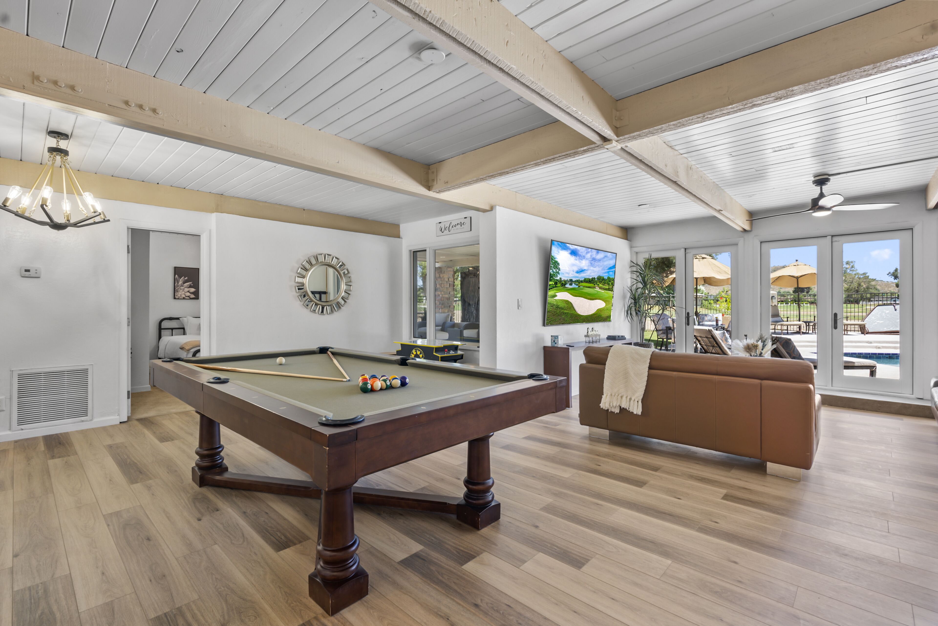 A pool table in a room with wooden floors, a brown sofa, and a TV displaying a golf course.