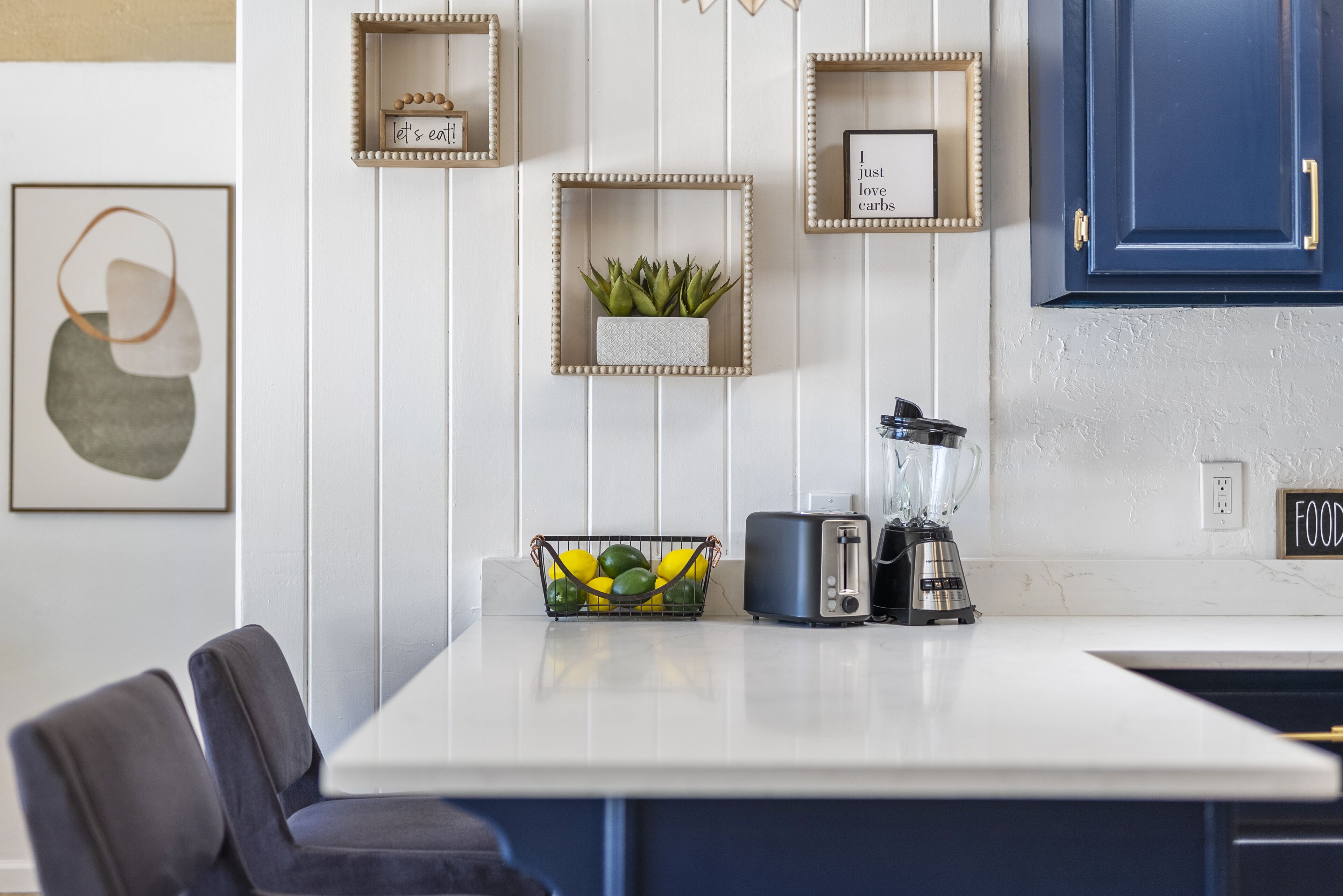 A kitchen with a white countertop, a toaster, a blender, and a fruit basket. There are framed pictures on the wall and a blue cabinet.
