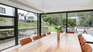 Een ruime eetruimte met een lange houten tafel en lederen stoelen, grote ramen met uitzicht op een tuin en omliggende huizen.