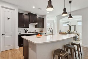 A modern kitchen with dark cabinets, a large island, pendant lighting, and a view into the dining area.