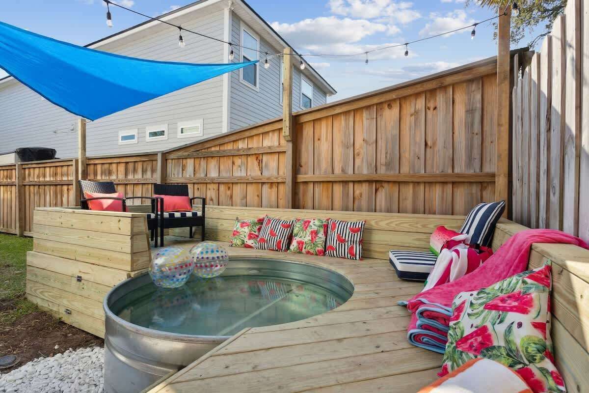 A backyard with a hot tub, wooden deck, outdoor furniture, and a blue shade sail.
