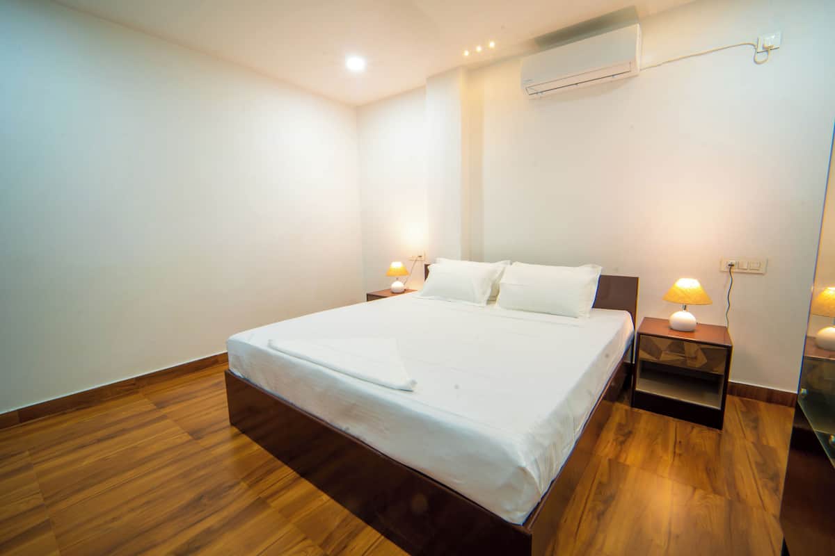 A neatly made bed in a hotel room with wooden flooring, a bedside table with a lamp, and an air conditioning unit on the wall.
