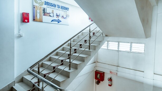 Una escalera con barandillas de metal que sube a un piso superior, un extintor en la pared, y un cartel publicitando un programa de residencia de té.