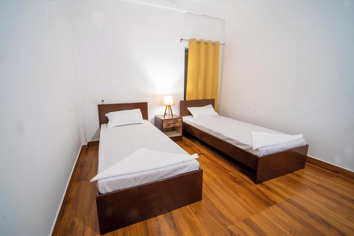 A room with two single beds, wooden flooring, a nightstand with a lamp, and a yellow curtain.