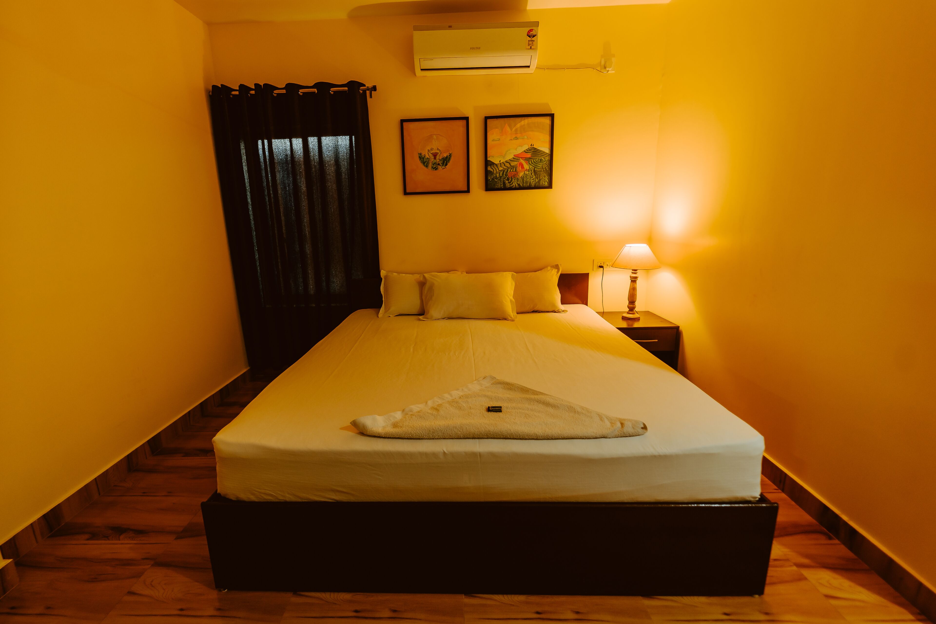 A narrow room with a single bed, a nightstand, a lamp, and two framed pictures on the wall.