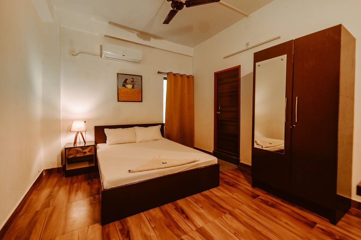 A bedroom with a bed, a nightstand, a ceiling fan, an air conditioning unit, a wardrobe, and a painting on the wall.