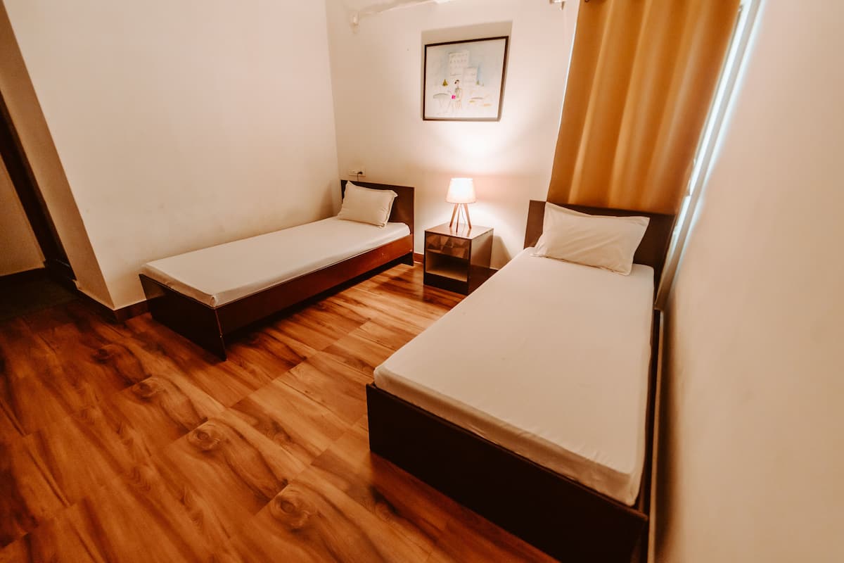 A room with two single beds, a nightstand with a lamp, wooden flooring, and a framed picture on the wall.
