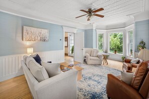 A spacious living room with a high ceiling, a ceiling fan, and large windows offering a view of the outdoors. - The Victorian - Reunite, Explore, Indulge! (Johnson)