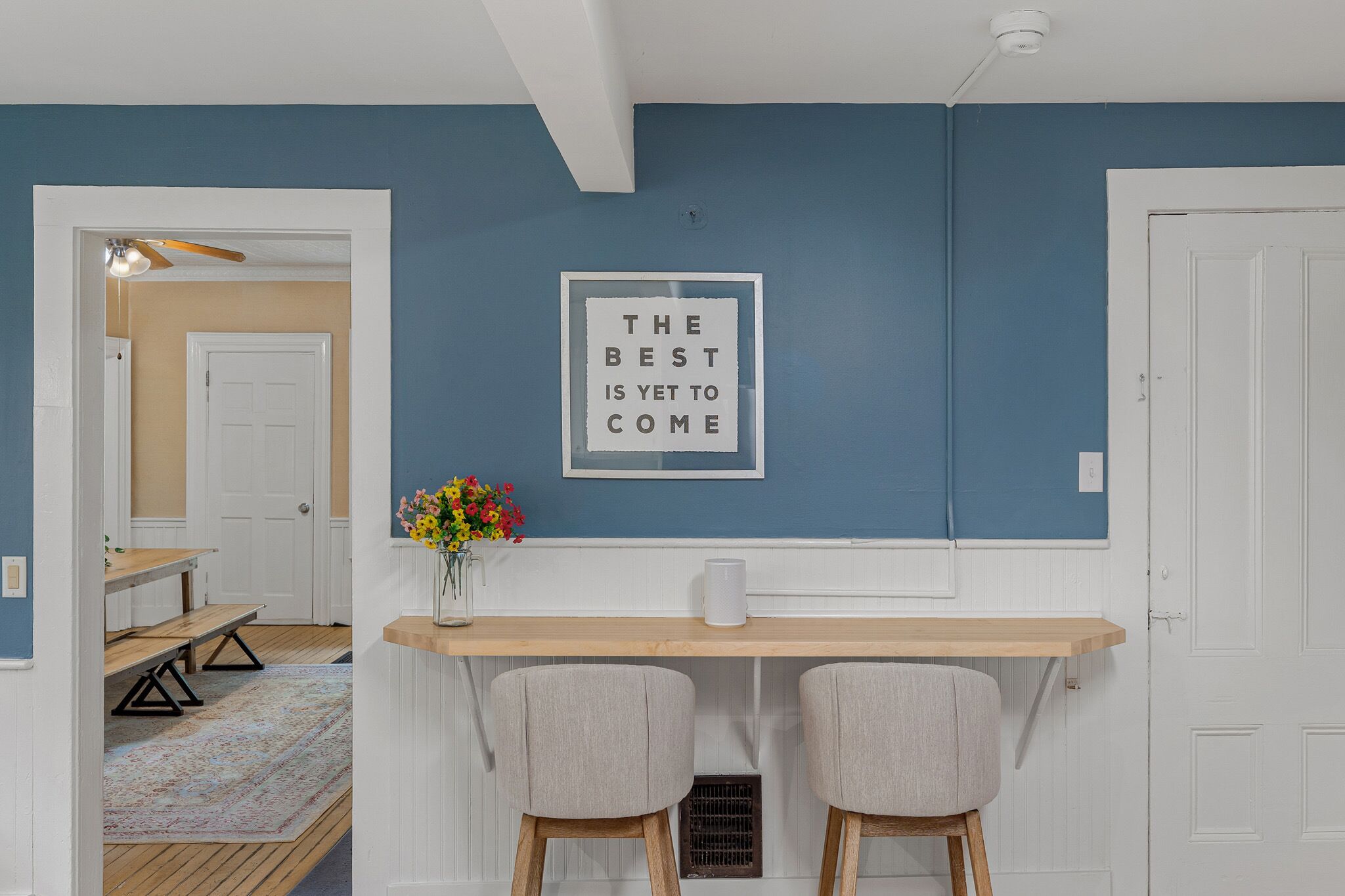 Une pièce avec un mur bleu, une porte blanche, une table en bois avec deux chaises et un cadre contenant une citation inspirante.