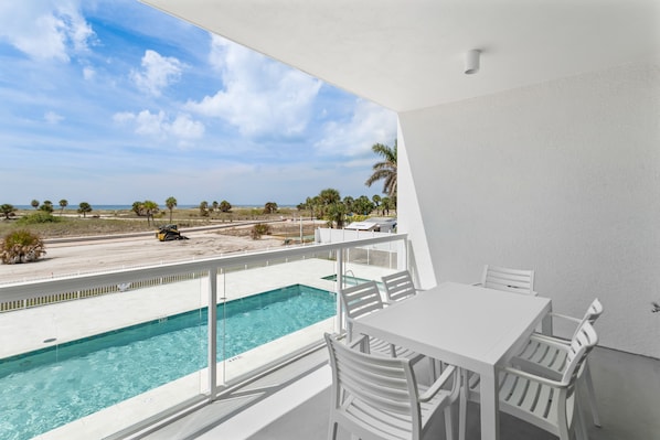 A balcony with a pool, white furniture, and a view of the beach and ocean.