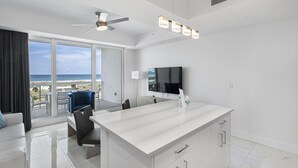 Una habitación de hotel moderna con un balcón grande, un ventilador de techo, un televisor de pantalla plana y una cocina blanca tipo isla.