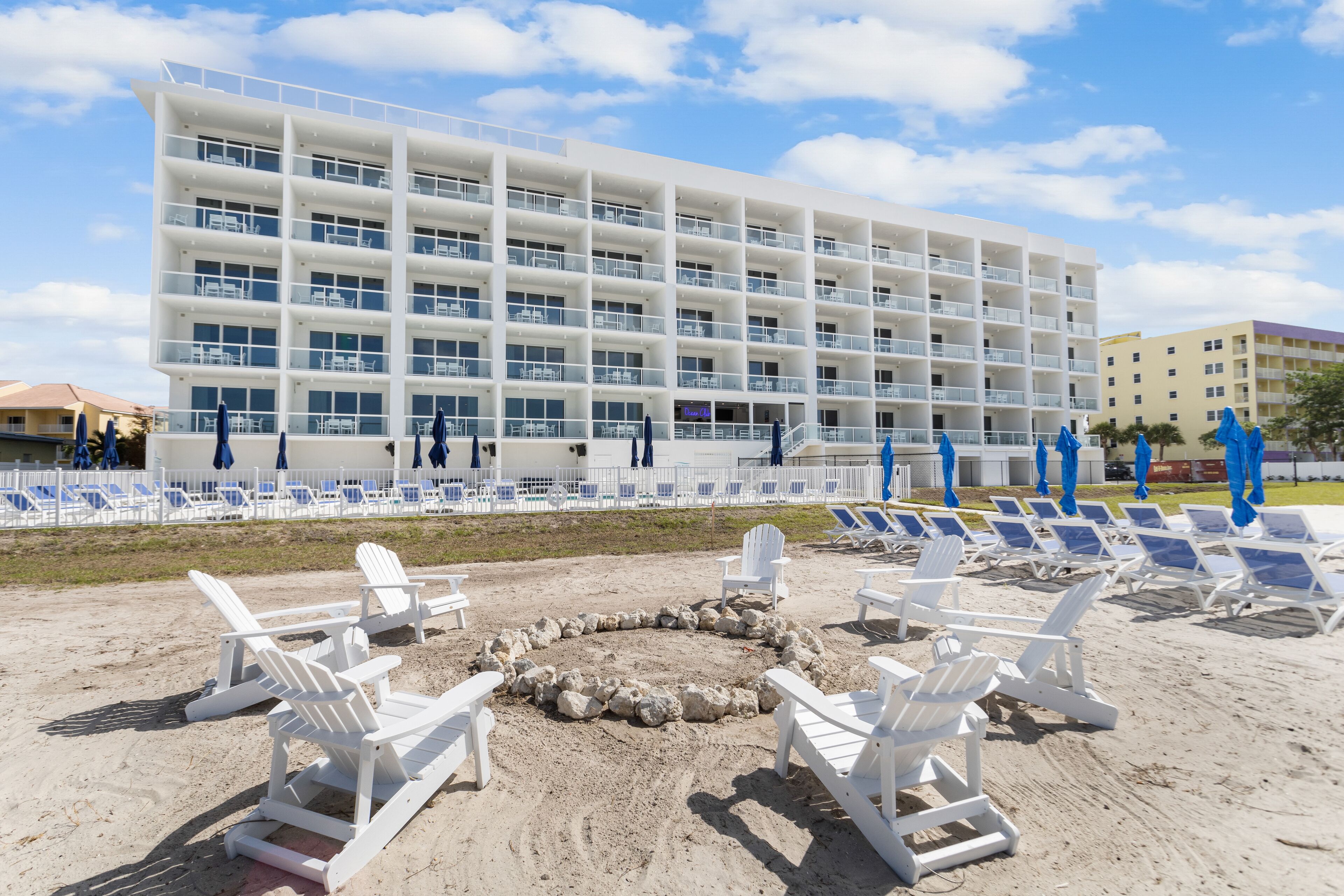 A beachfront hotel with multiple balconies, a sandy beach area with lounge chairs, and a large building with a white facade.