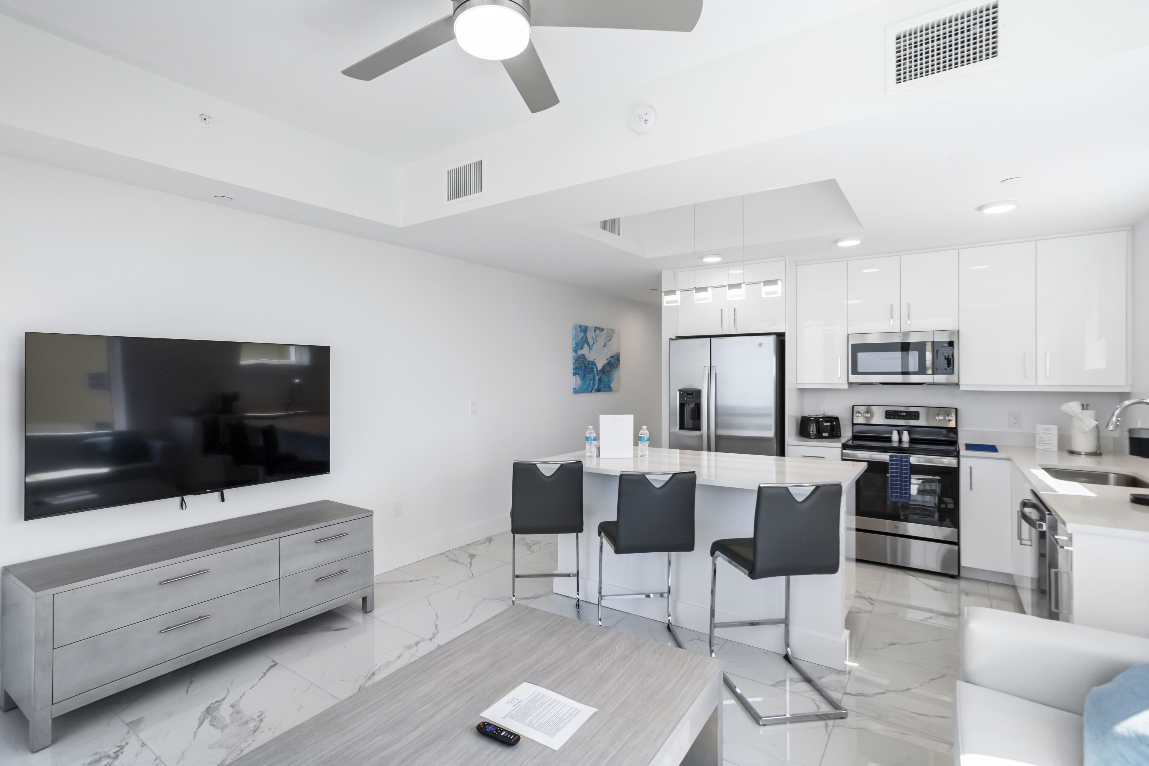Una cocina moderna con armarios blancos, electrodomésticos de acero inoxidable y un televisor de pantalla plana grande. Hay una zona de comedor con encimera de mármol y sillas negras.