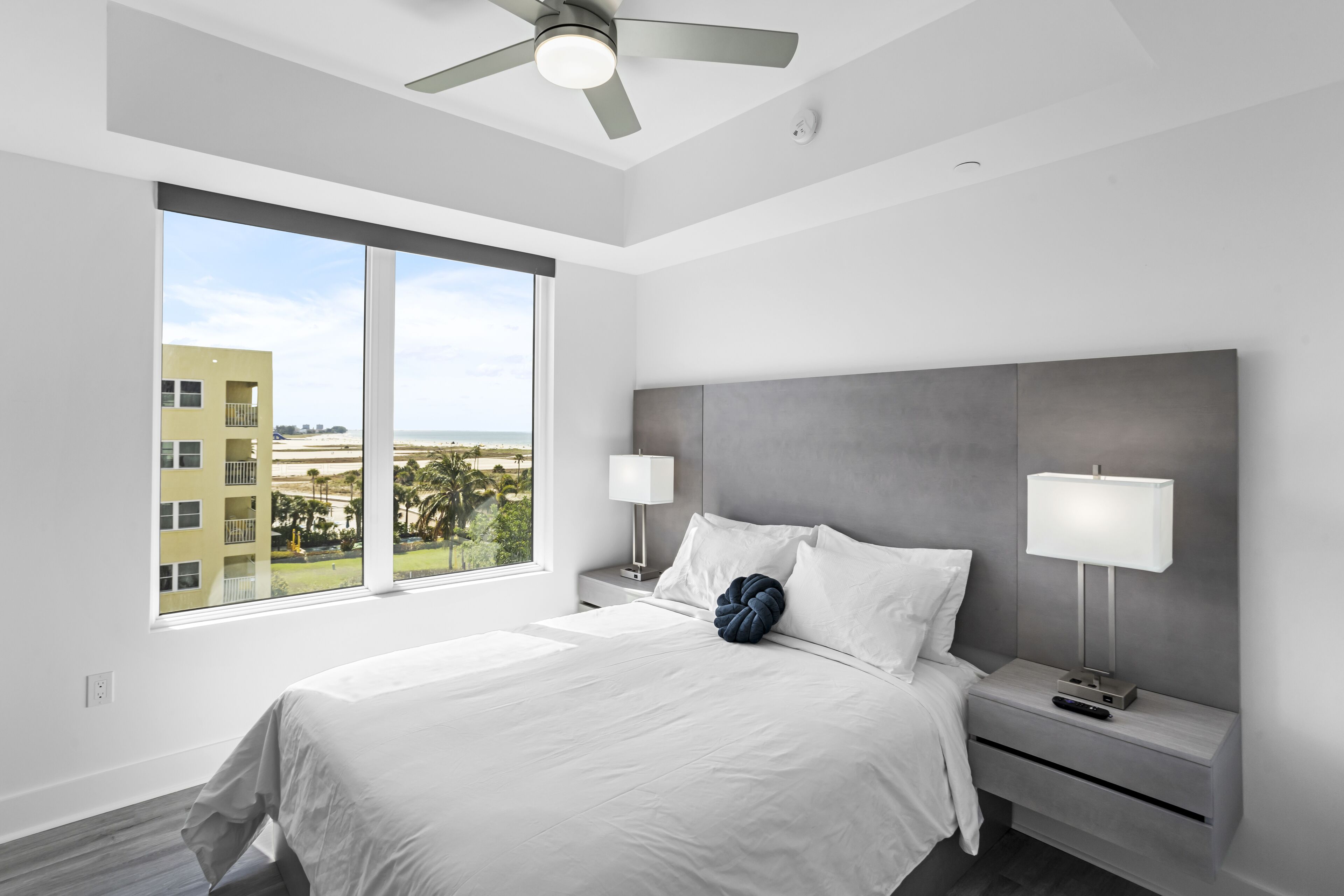 A modern hotel room with a large bed, two bedside tables, a ceiling fan, and a view of the beach.