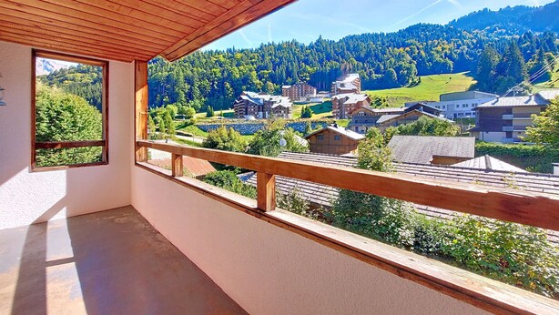 Un balcon doté d’une balustrade en bois, donnant sur un village aux bâtiments traditionnels et sur une colline boisée.