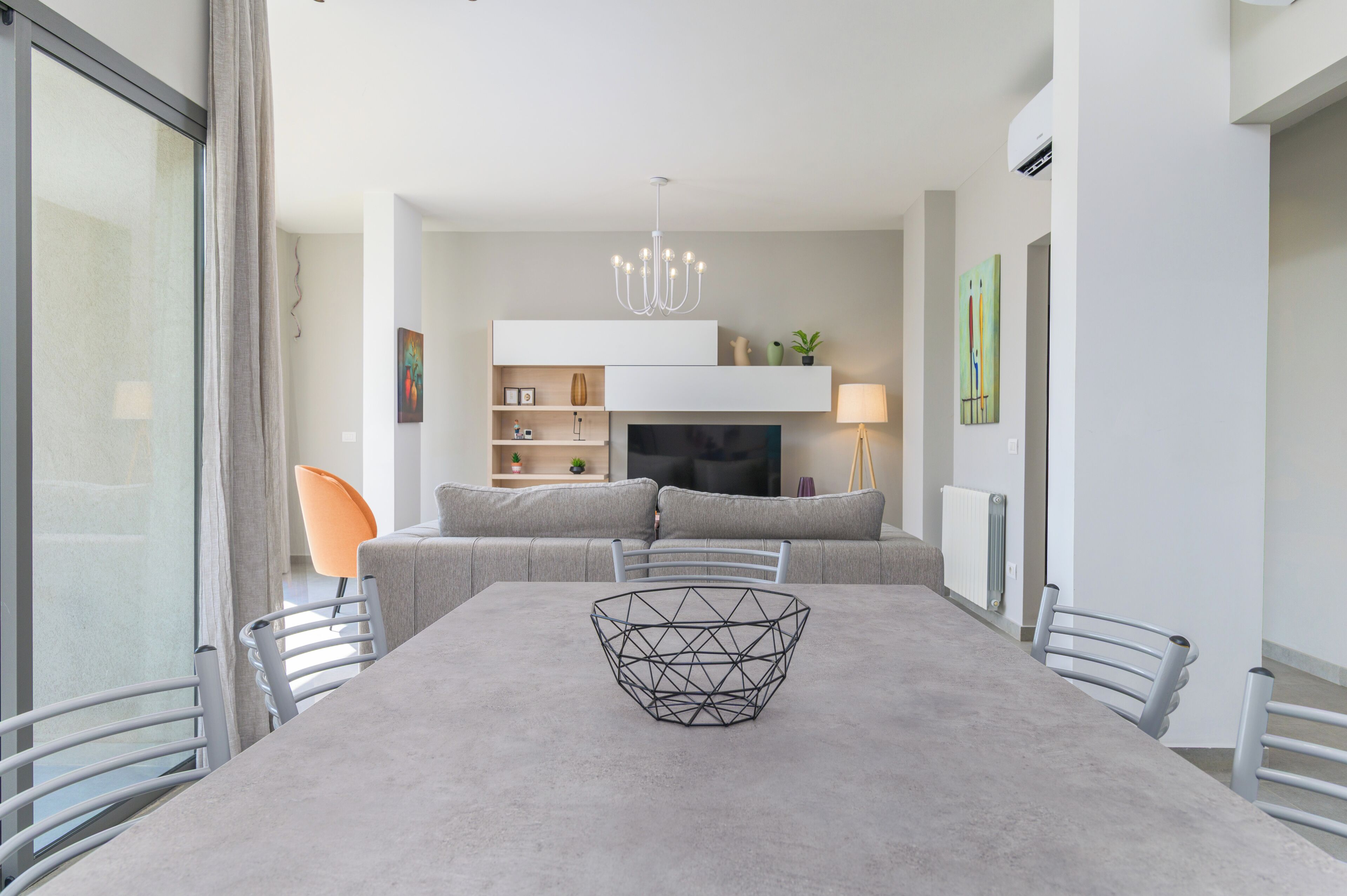 A modern dining area with a large table, chairs, a geometric centerpiece, a sofa set, a TV, and a chandelier.
