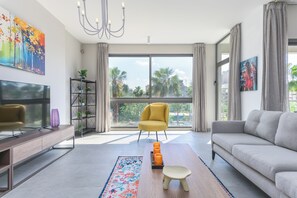 A modern living room with a sofa, a yellow armchair, a TV, a bookshelf, and a large window with a view of palm trees. - Le 115 Aparthotel in Jal El Dib (Jal El Dib)