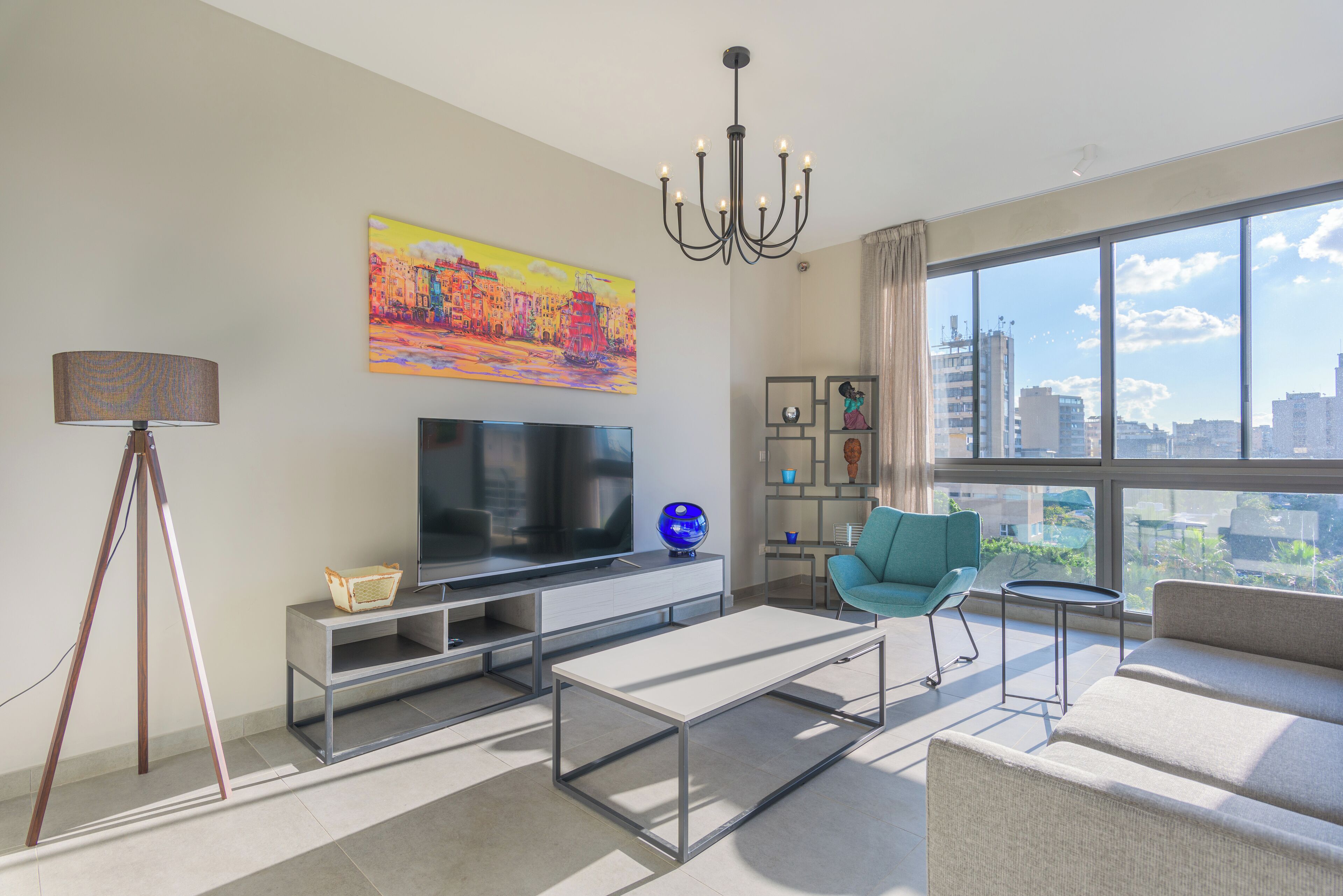 A modern living room with a large window, a flat-screen TV, a blue chair, and a chandelier.