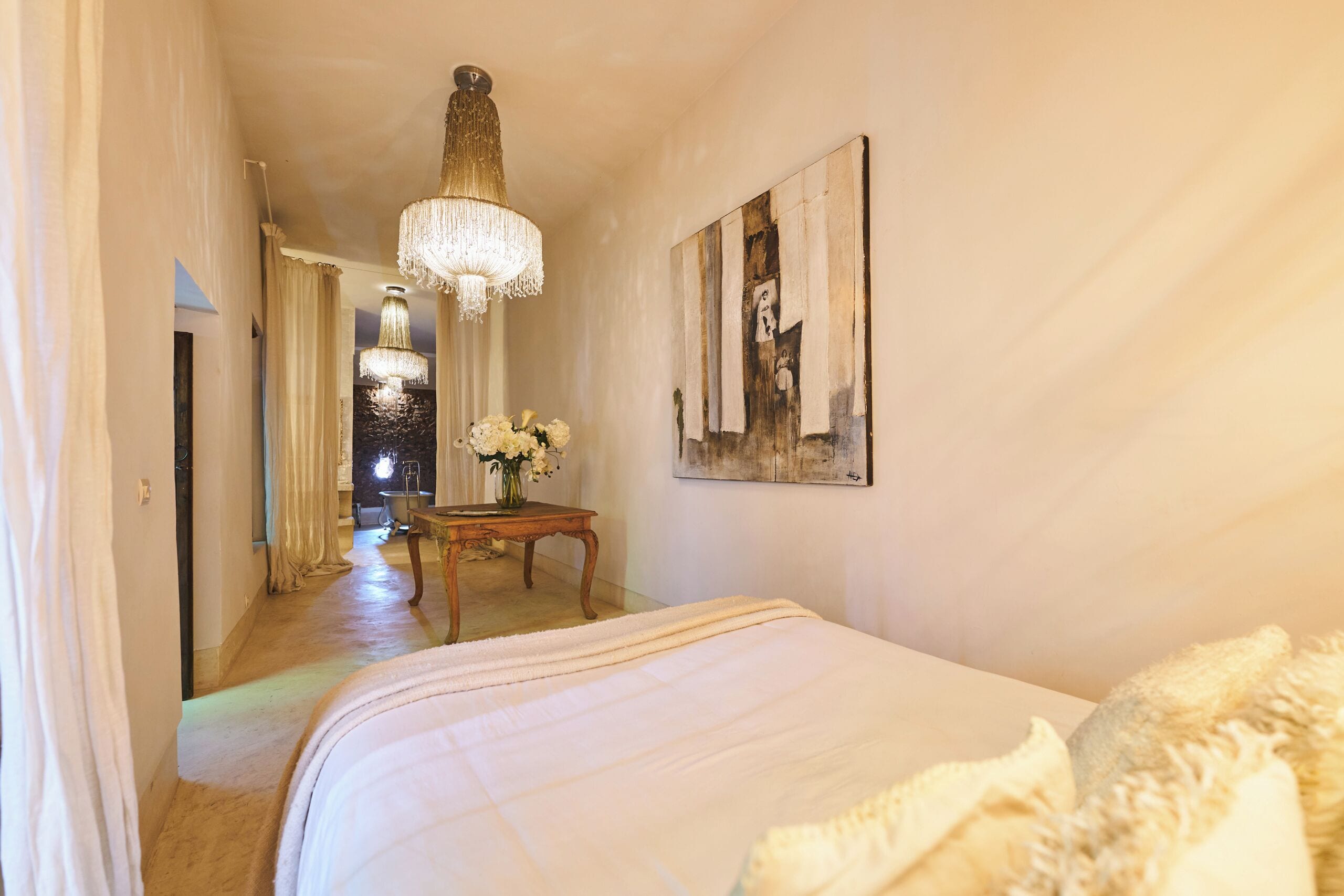 A hotel room with a large bed, a chandelier, a wooden table with flowers, and a painting on the wall.
