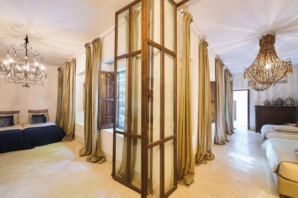 A room with two beds, a chandelier, and a large window with curtains.