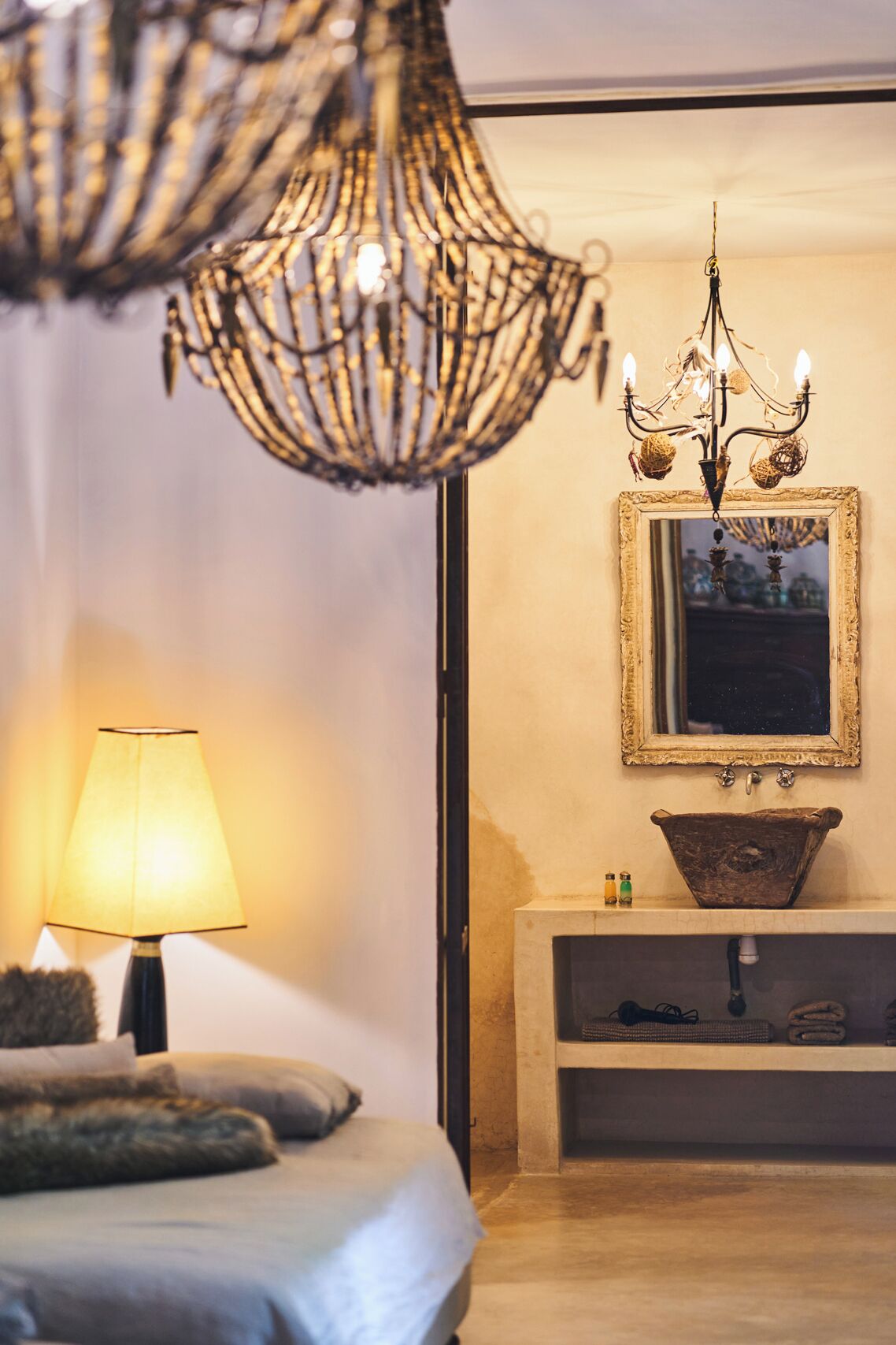 A bedroom with a bed, a bedside lamp, a chandelier, a mirror, and a wooden console table with a bowl.