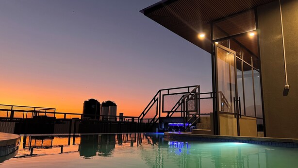 Un área de piscina con vista al atardecer, una zona de estar cubierta y una escalera que conduce a un nivel superior.