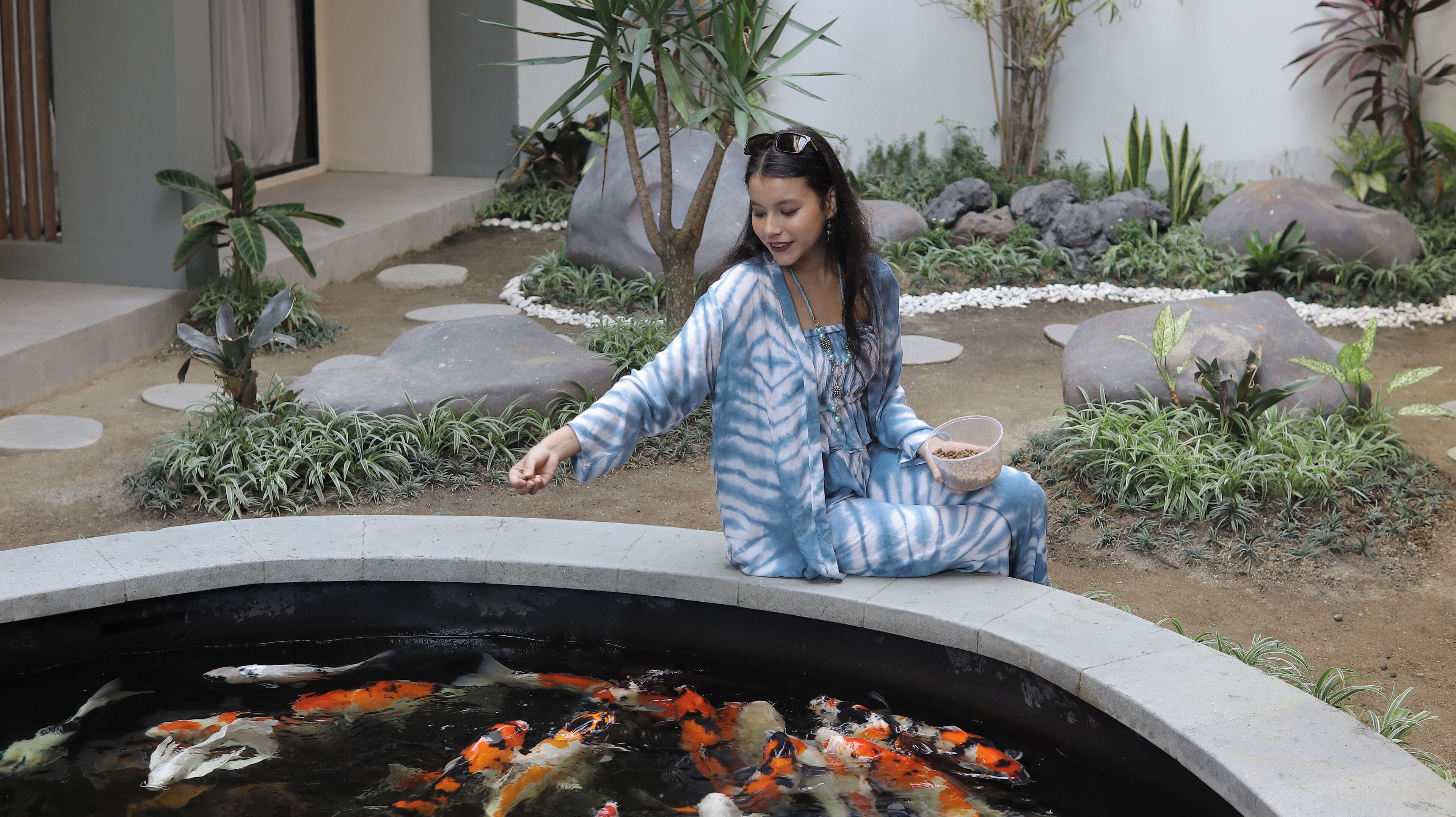 A woman sitting by a koi pond in a garden with rocks and plants.