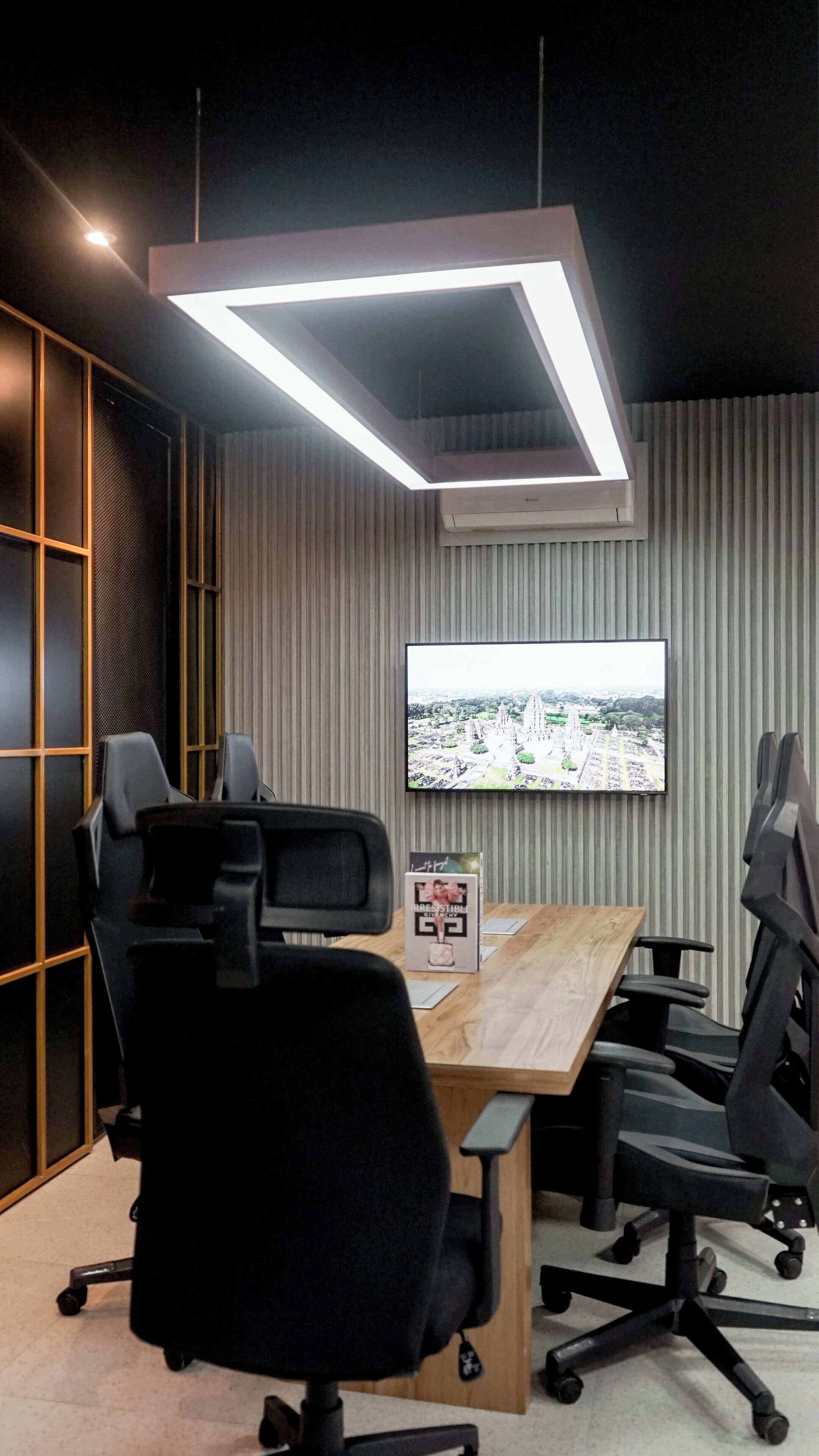 A modern conference room with a large table, black office chairs, a mounted TV displaying a cityscape, and a unique ceiling light fixture.