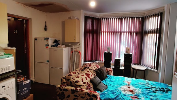 A compact hotel room with a bed, a small desk, a chair, a washing machine, and a ceiling projector.